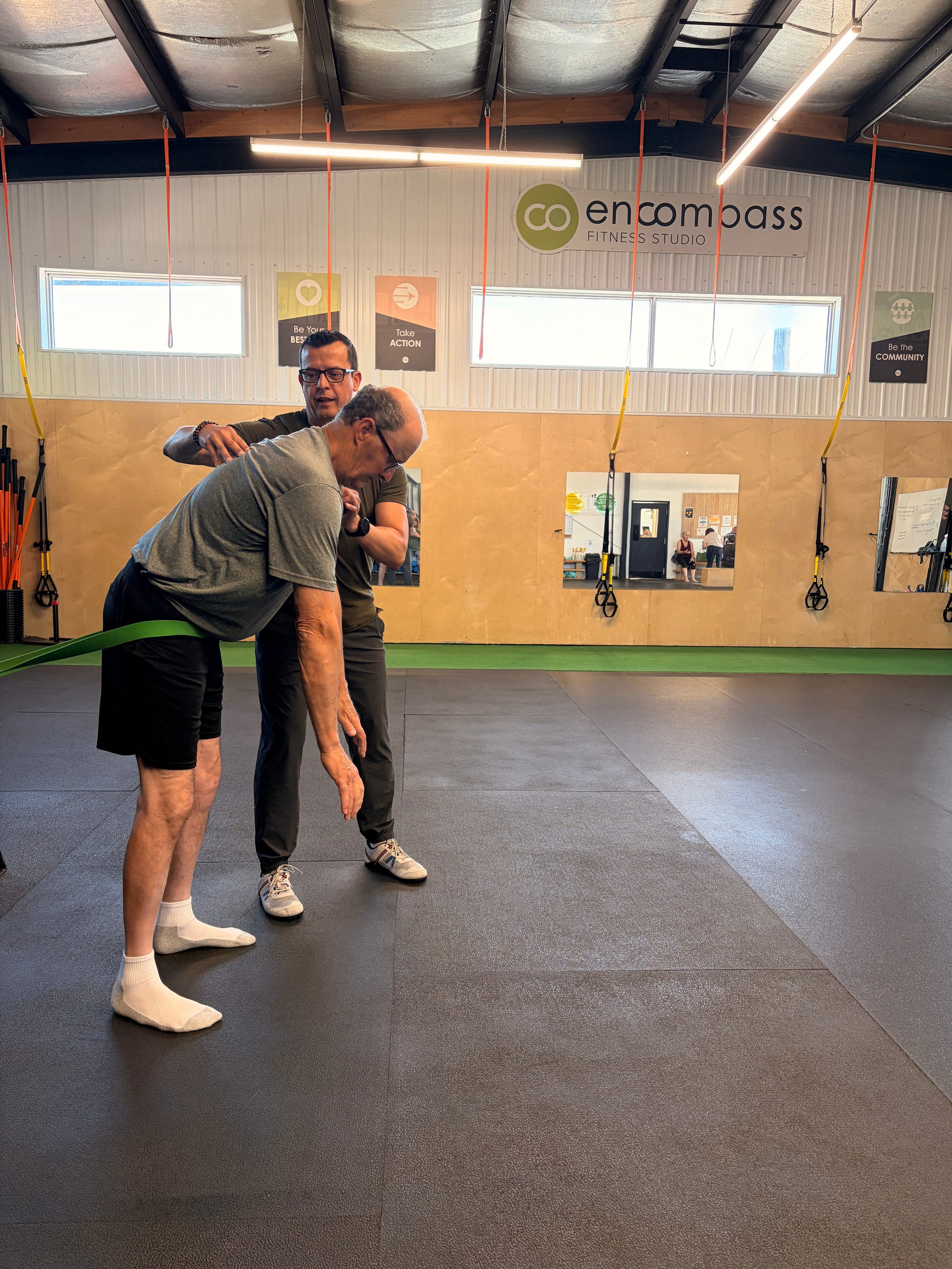 Two men in a gym, one an older man with glasses and a gray t-shirt, bending forward, the other a younger man with glasses and a dark t-shirt, assisting him with training. The gym has black rubber flooring and walls with motivational posters, featuring a large window and mirrors.