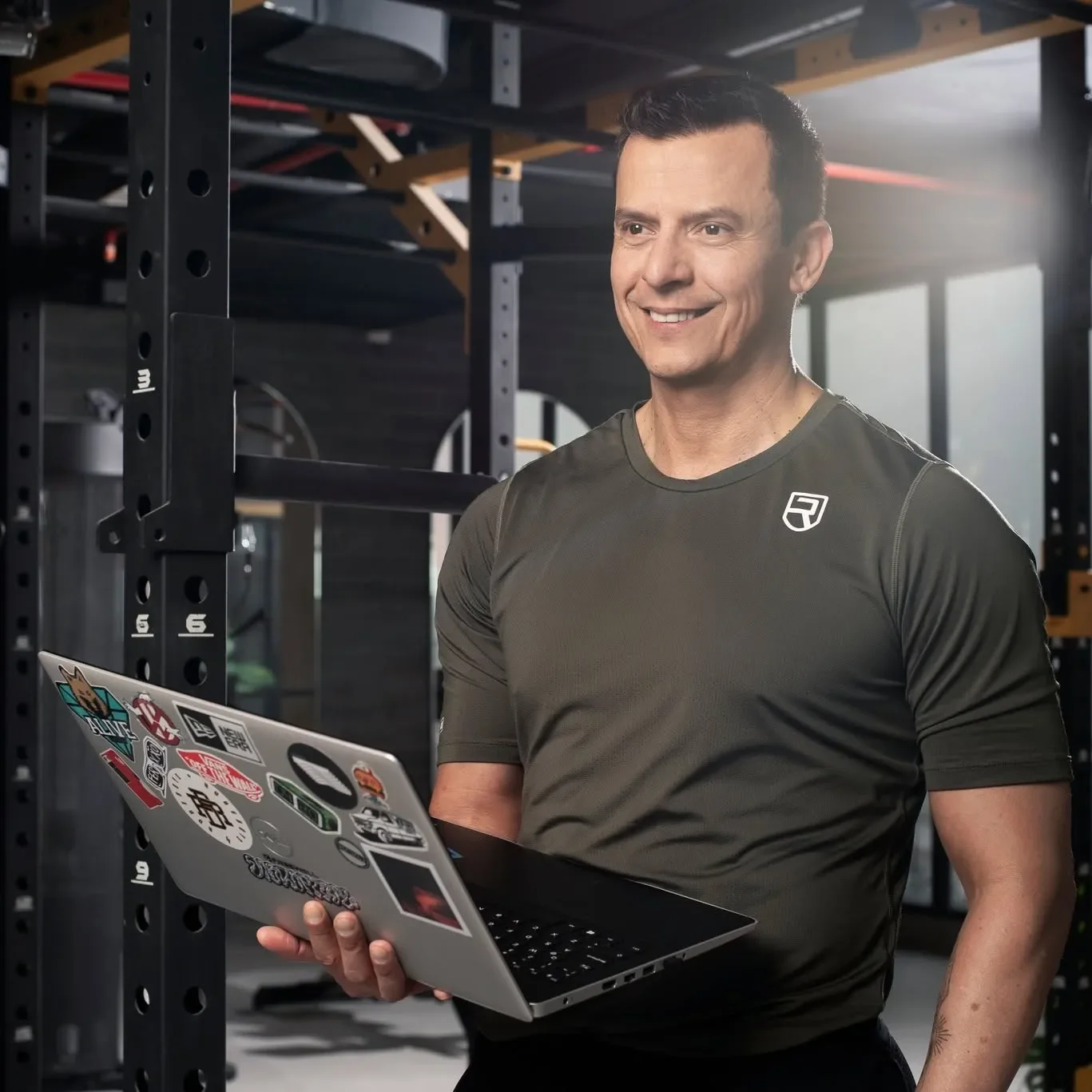 A man in athletic clothing holds a laptop covered in various stickers in a gym setting with gym equipment and large windows in the background.