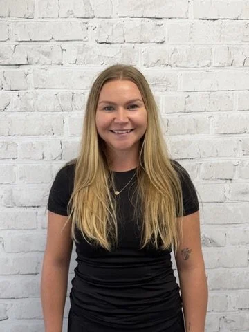A woman with long blonde hair smiling, standing against a white brick wall, wearing a black t-shirt.