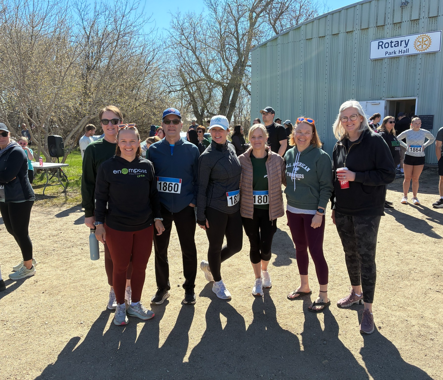 Group of people standing together outdoors near a building with a sign that reads "Rotary Park Hall." The group includes men and women of various ages, some wearing race bibs, athletic clothing, and sunglasses, indicating they are participating in a running or walking event on a sunny day.