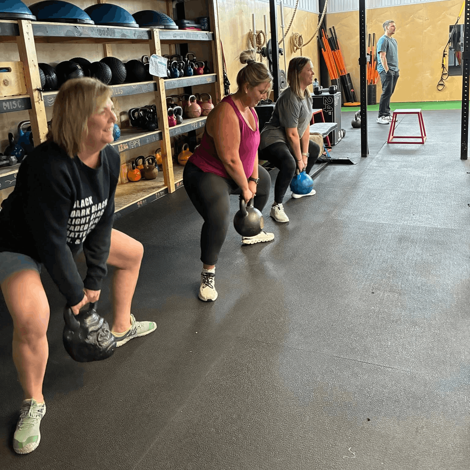 Four women performing kettlebell exercises in a gym, with a man in the background standing near a wall with orange workout sticks and ropes.