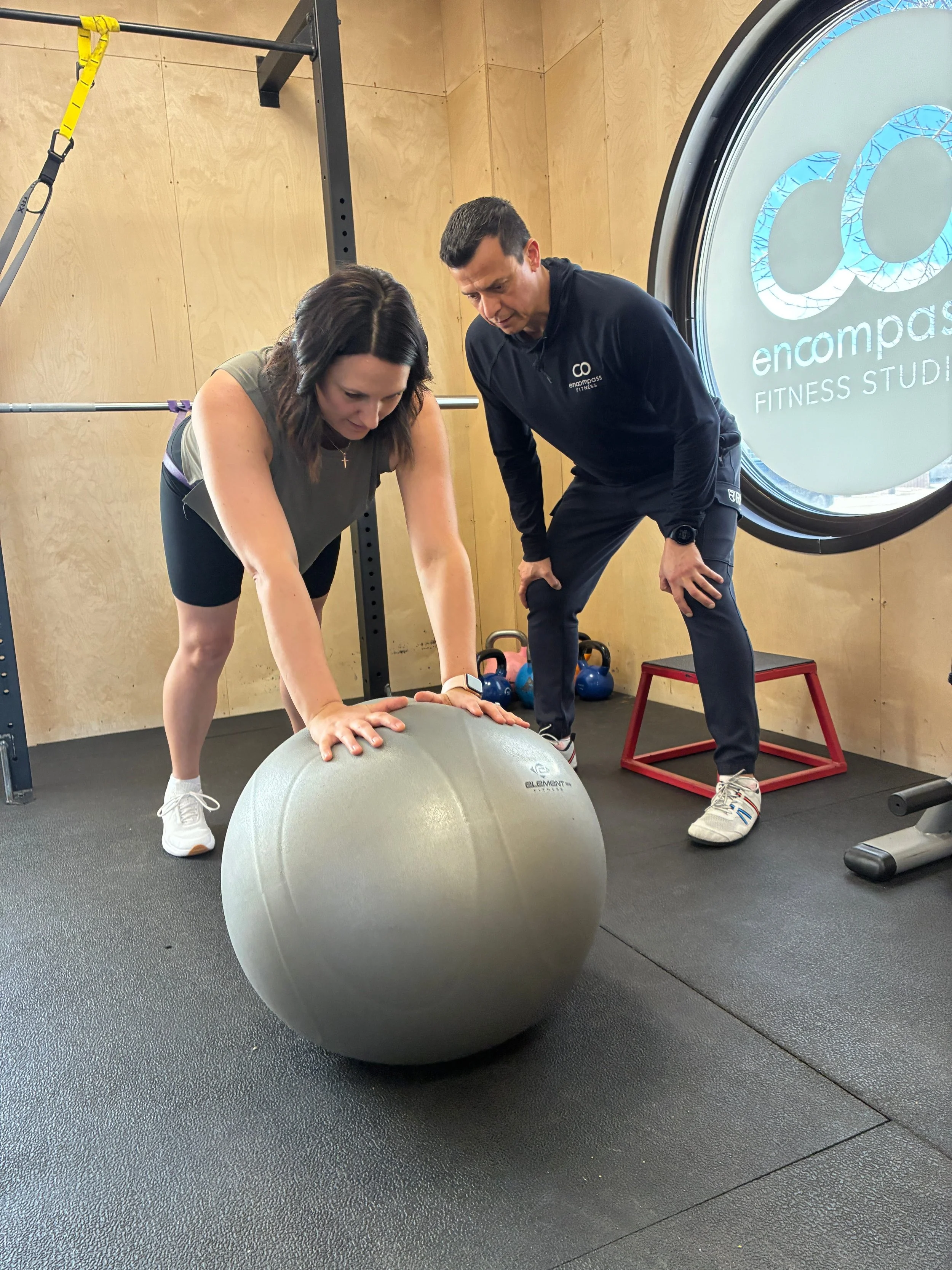 A woman and a man are in a gym, with the woman doing a plank exercise on a large exercise ball and the man acting as her trainer, leaning forward, observing her form.