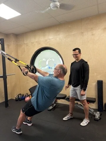 A man in a blue shirt and black shorts exercises with TRX suspension straps in a gym, while another man in a black shirt and gray shorts observes.