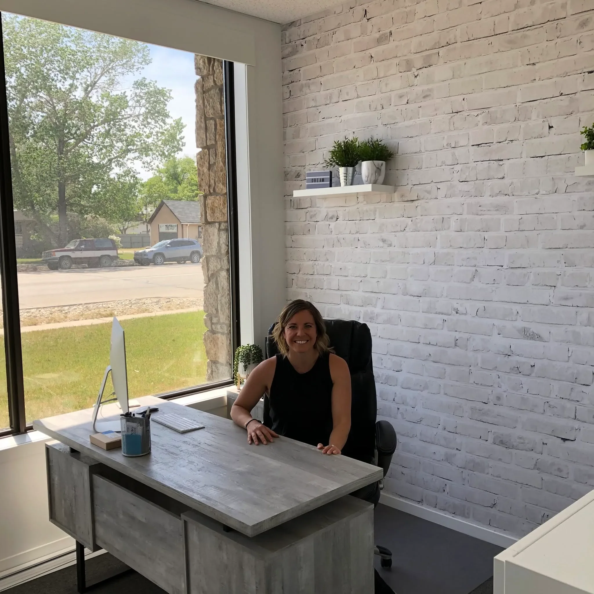 Woman sitting at a desk in a modern office with a white brick wall and large window showing trees and parked cars outside.
