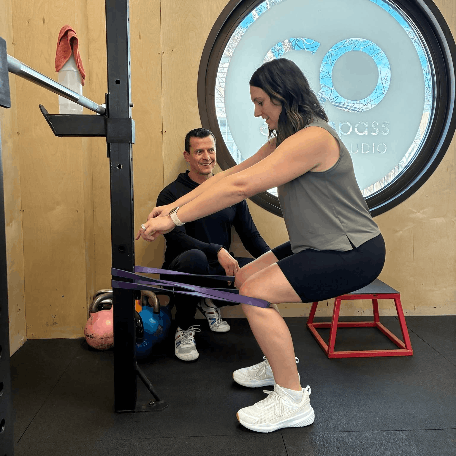 Woman in workout attire squatting with resistance band in gym, man seated on the floor smiling, kettlebells and gym equipment nearby, window with logo in background.