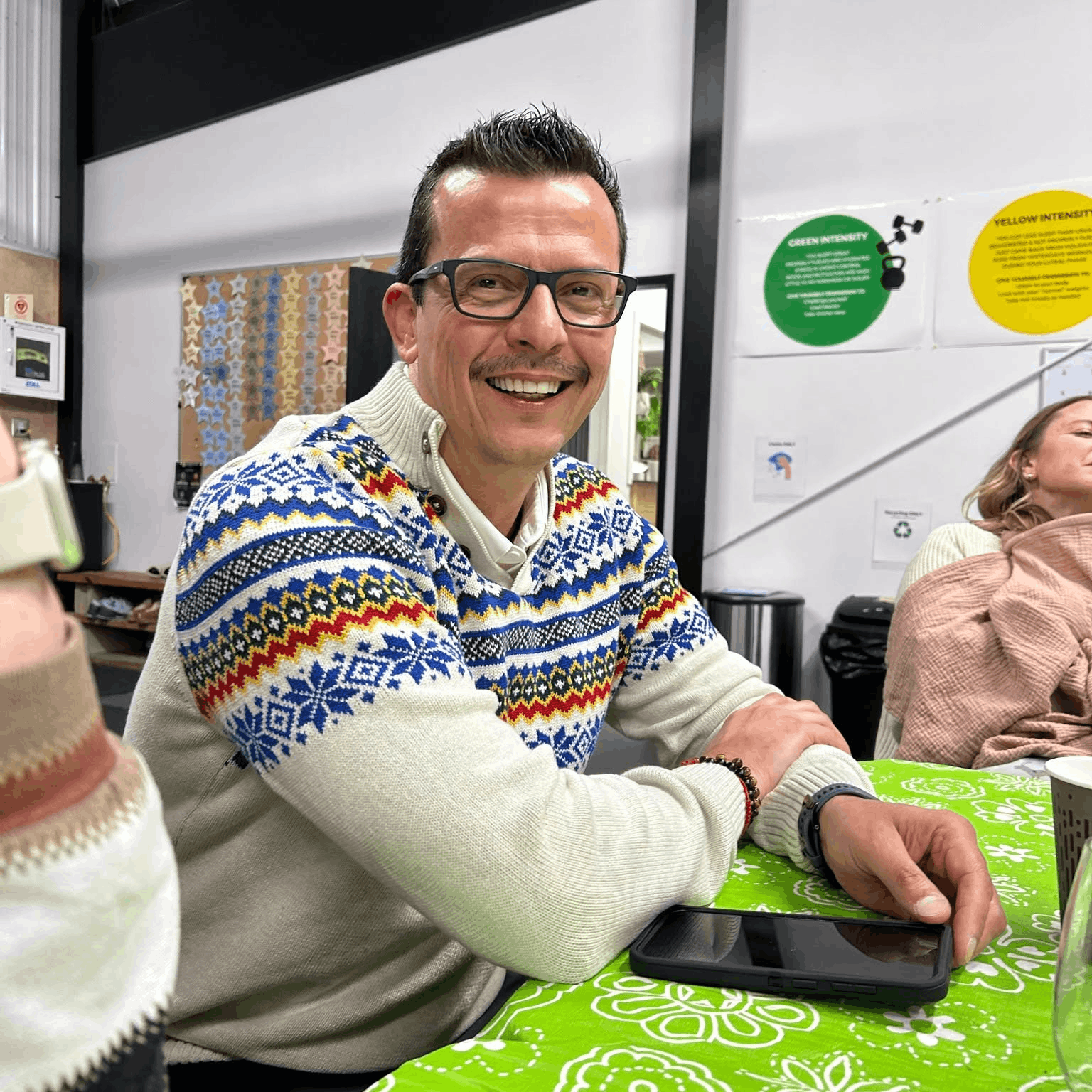 A man with glasses smiling at a table decorated with a green tablecloth, wearing a colorful holiday sweater, with a woman sitting next to him and a phone on the table.