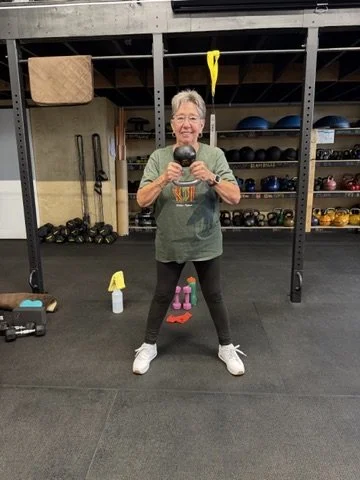 Older woman smiling and taking a selfie with a kettlebell in a gym, surrounded by fitness equipment including dumbbells, kettlebells, and medicine balls.