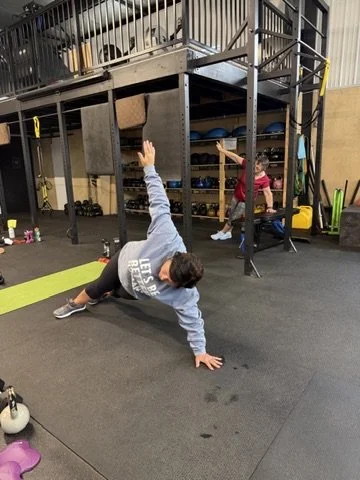 A man doing a plank exercise on the floor inside a gym with kids in the background.