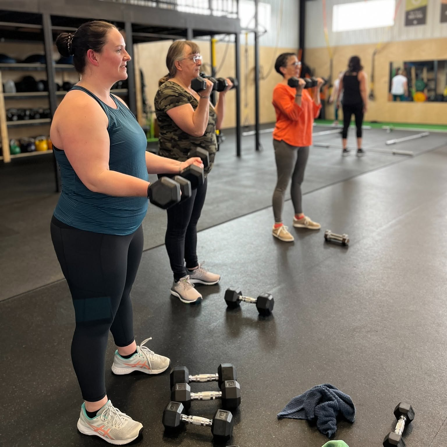Women working out in a gym lifting dumbbells, with equipment shelves and other gym members in the background.