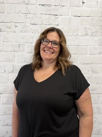 A woman with shoulder-length brown hair and glasses, wearing a black shirt, smiling in front of a white brick wall.