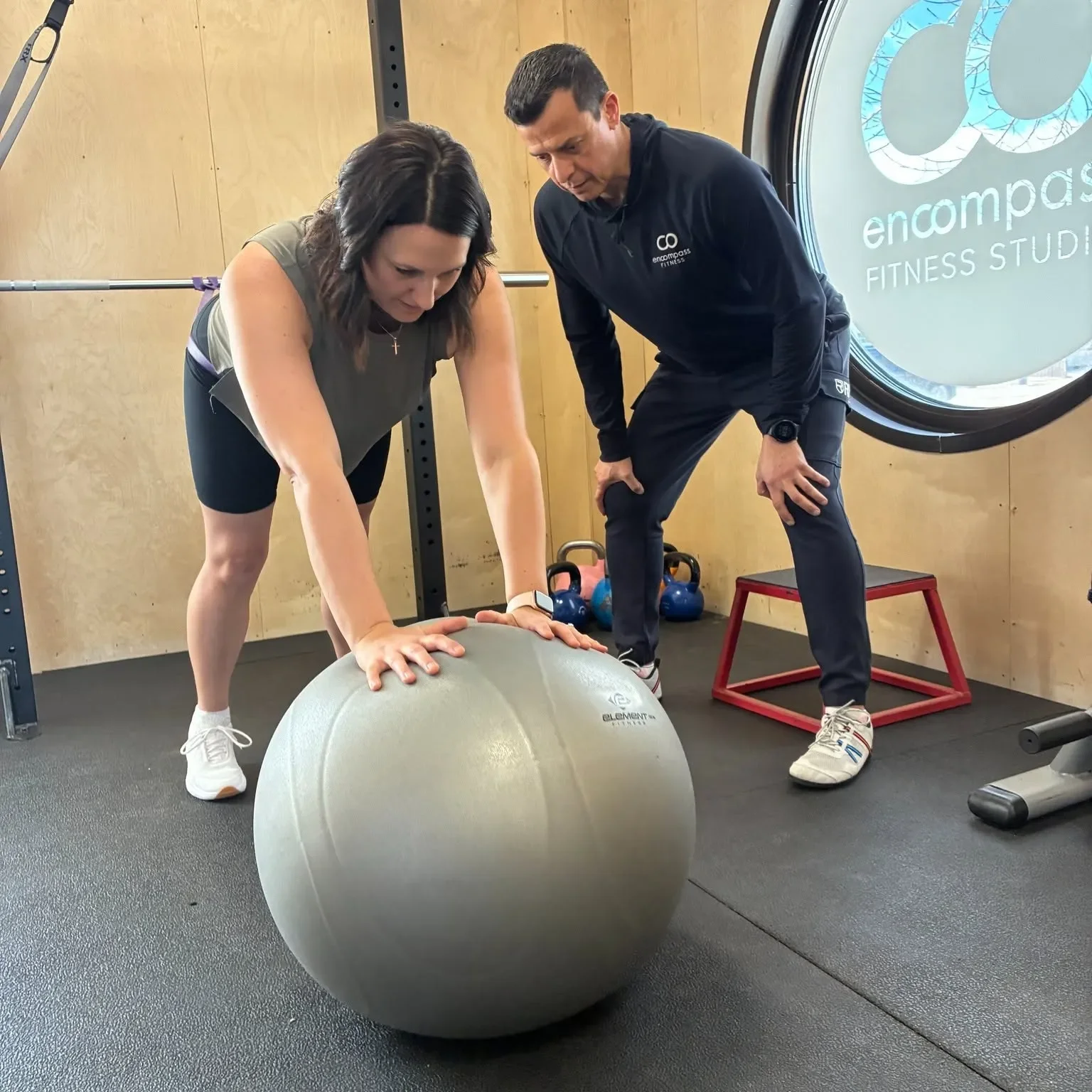 A woman and a man are doing a fitness exercise with a large exercise ball in a gym. The woman is leaning forward on the ball with her hands on it, and the man is standing next to her, watching and instructing.