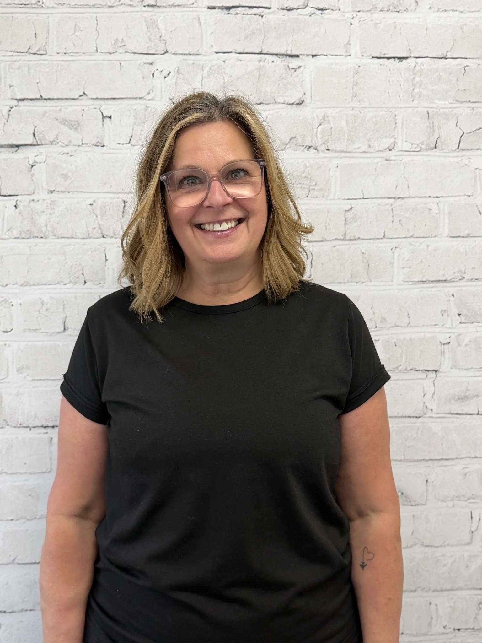 A woman with shoulder-length blonde hair, wearing glasses and a black t-shirt, smiling and standing against a white brick wall.