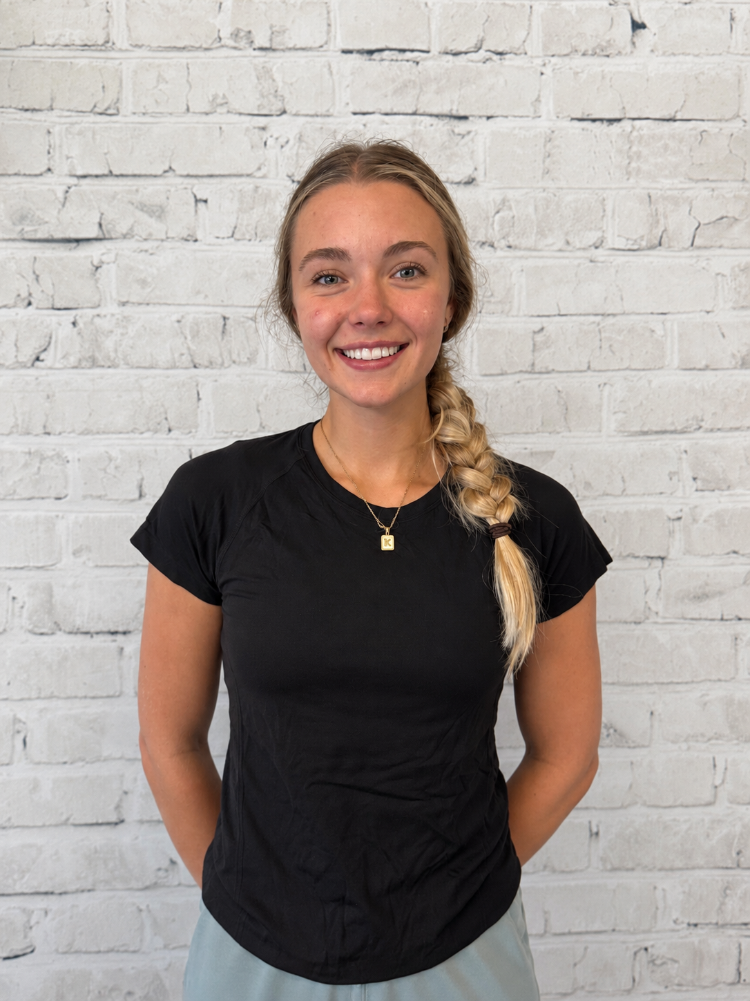 A young woman with blonde hair in a braid, smiling, wearing a black t-shirt and a necklace with a gold pendant, standing against a white brick wall.