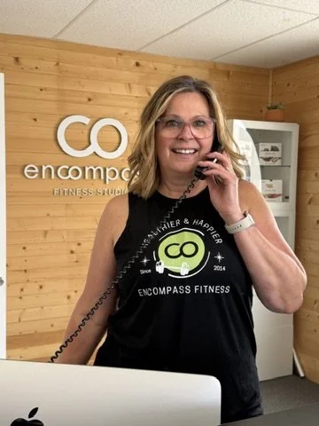 Woman taking a selfie in a fitness studio with a wooden wall, wearing a black tank top with sports logo, smiling, with a phone in one hand and an Apple computer in front.