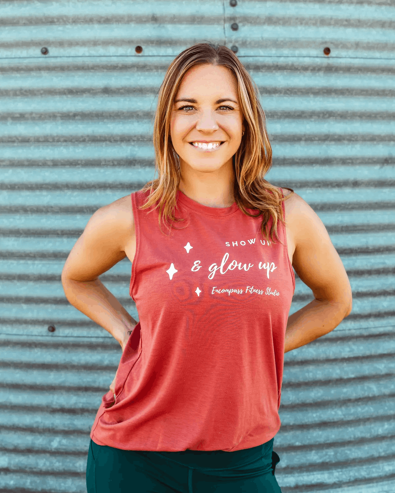 A woman with shoulder-length brown hair wearing a red sleeveless shirt and black pants standing in front of a corrugated metal wall, smiling with hands on hips.