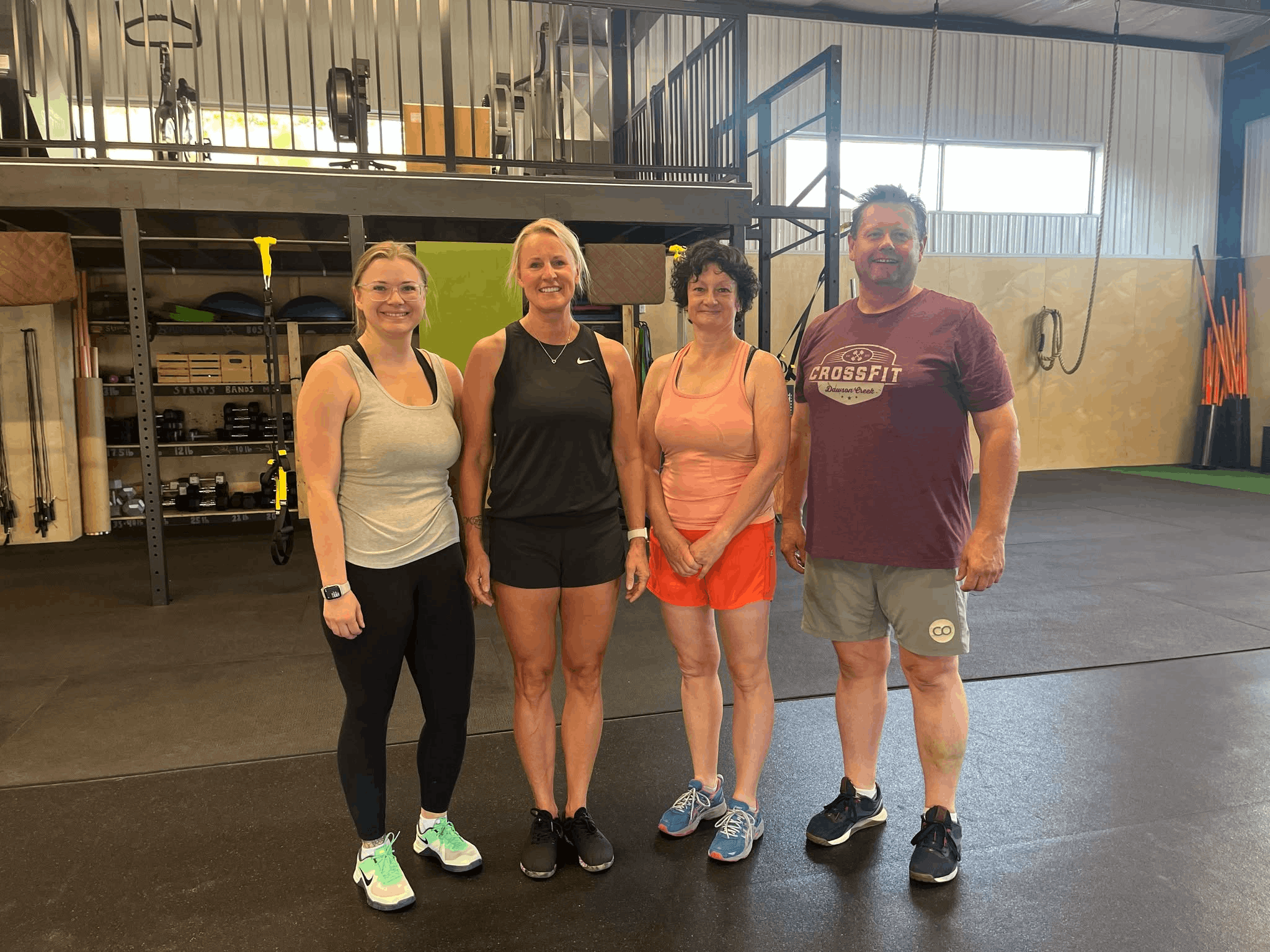 Four people standing inside a gym, smiling at the camera. They are dressed in workout clothes, and gym equipment such as weights and resistance bands are visible in the background.