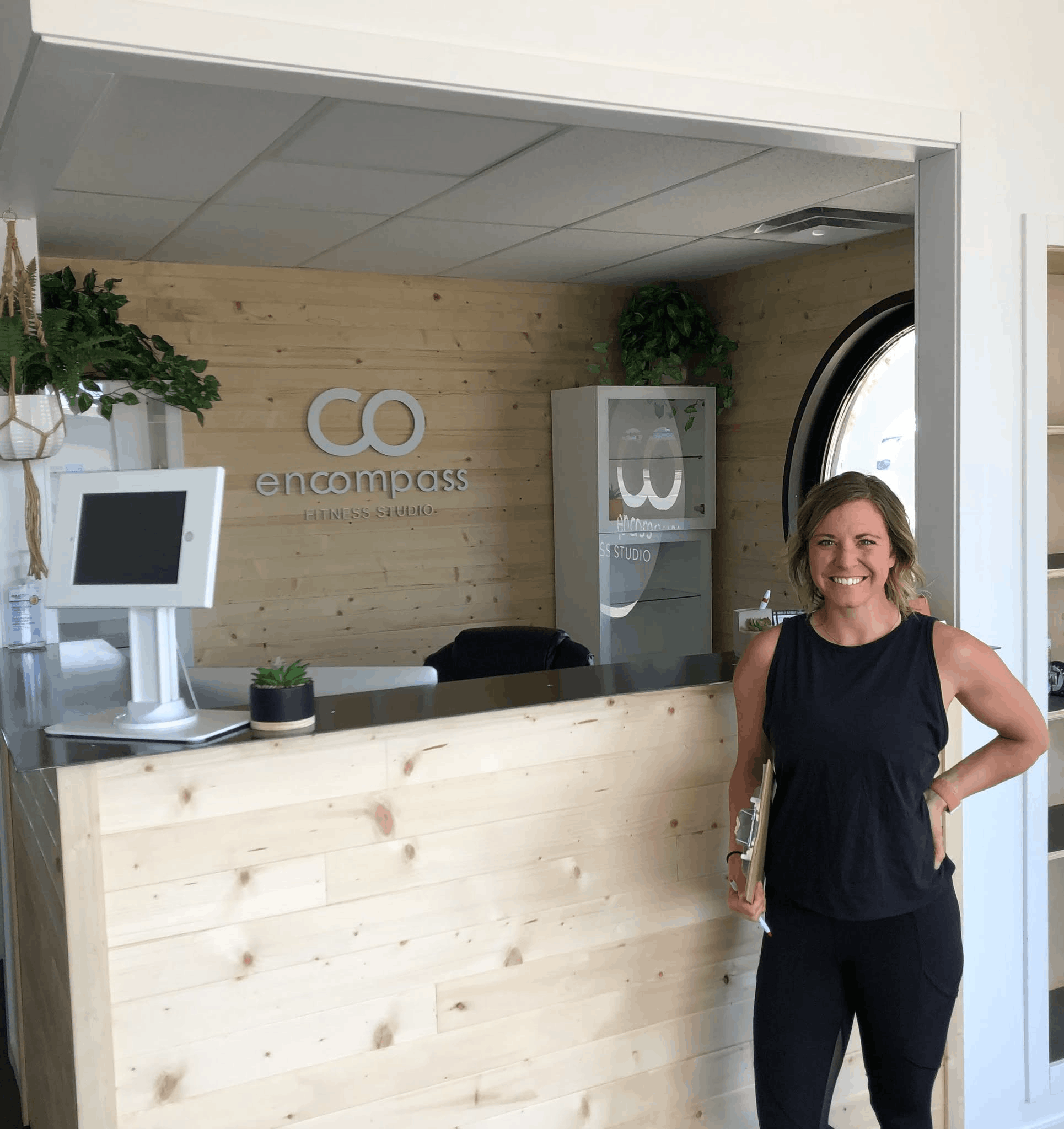 Woman with short blonde hair smiling in front of a wooden wall with plants and a sign that reads 'co compass fitness studio'.