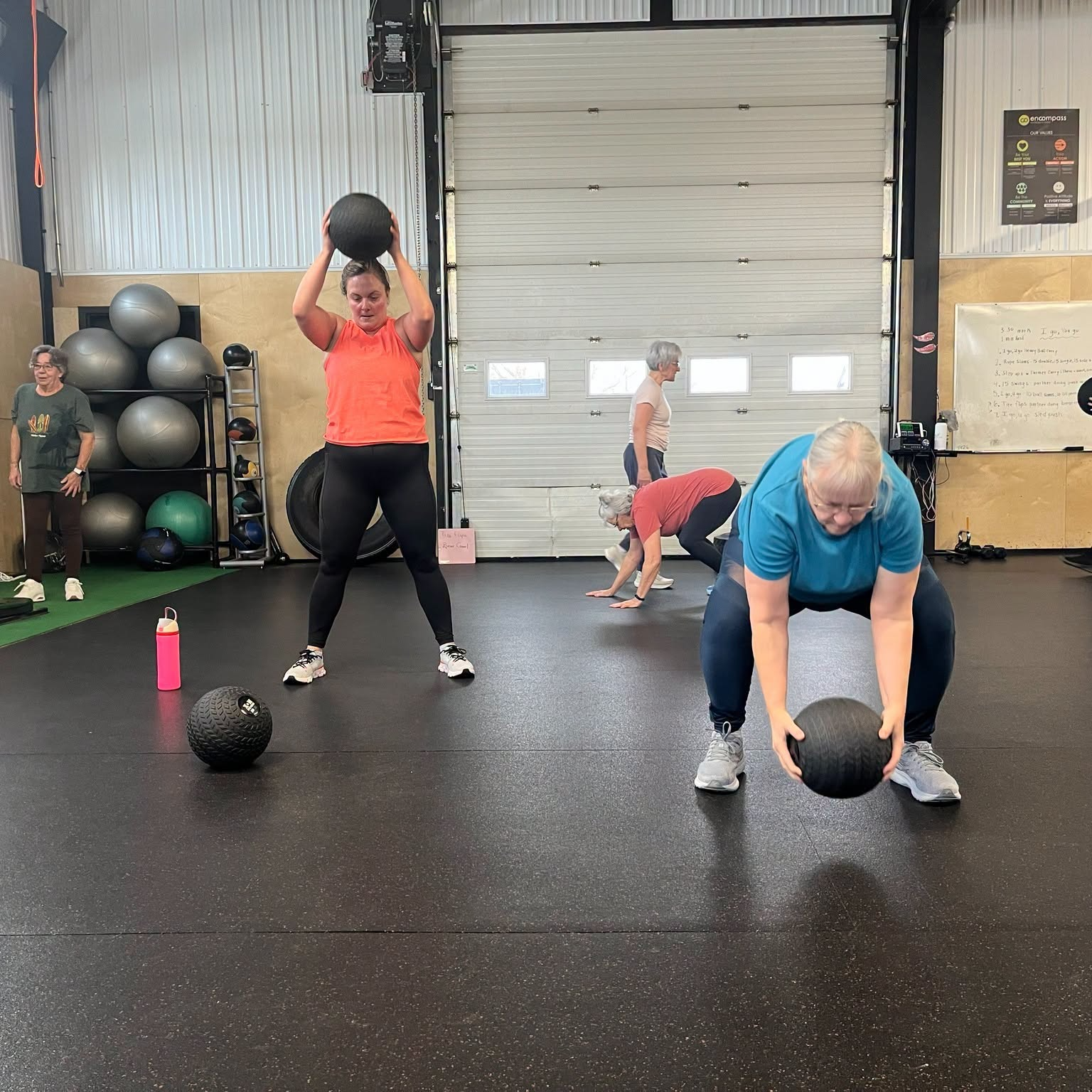 Several women working out in a gym, performing exercises with medicine balls and a farmer's walk with a weighted ball, under the supervision of a trainer.