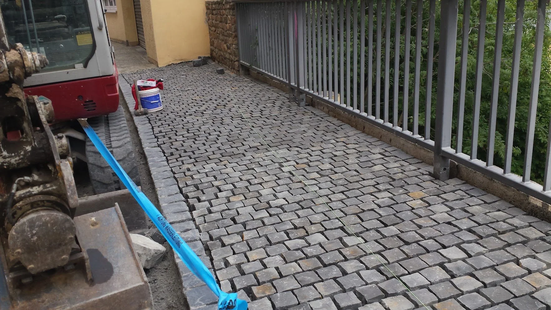 Blick auf eine mit Kopfsteinpflaster ausgelegte Terrasse oder Gehweg, der im Bau ist. Es sind Bauwerkzeuge und -materialien, darunter ein Mörtelbecher, eine Wasserwaage, Steine und eine Schnur, sichtbar. Ein Zaun und eine Gebäudefassade sind im Hinte