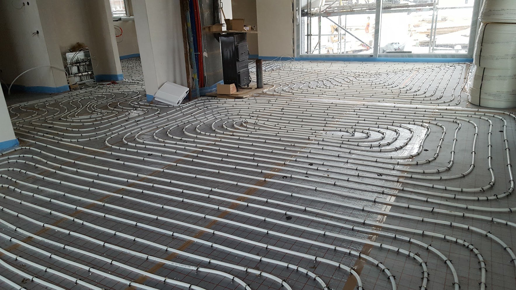 Installationsarbeit an einer Fußbodenheizung mit sichtbaren weißen Rohrleitungen, die in einer Musterung installiert sind, in einem Raum unter Bauarbeiten, mit großen Fenstern im Hintergrund.