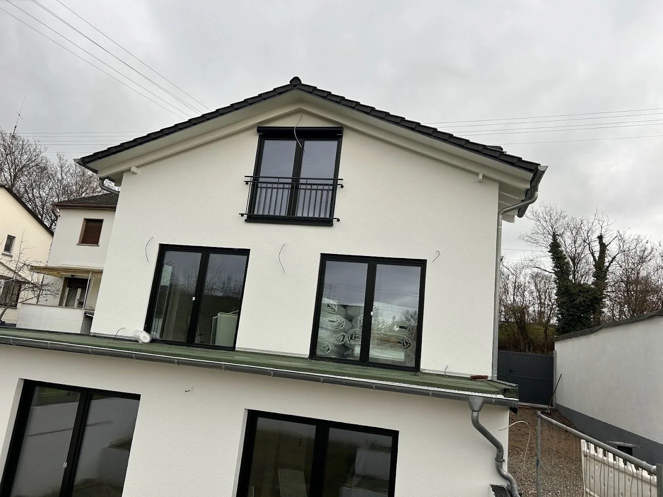 Neues zweistöckiges Haus mit weißen Fassaden, großen Fenstern und schwarzen Rahmen, Dach mit Ziegeln, Dächerschäden sichtbar, Gully mit Rohrleitung, umgeben von Bäumen und Nachbarhäusern.