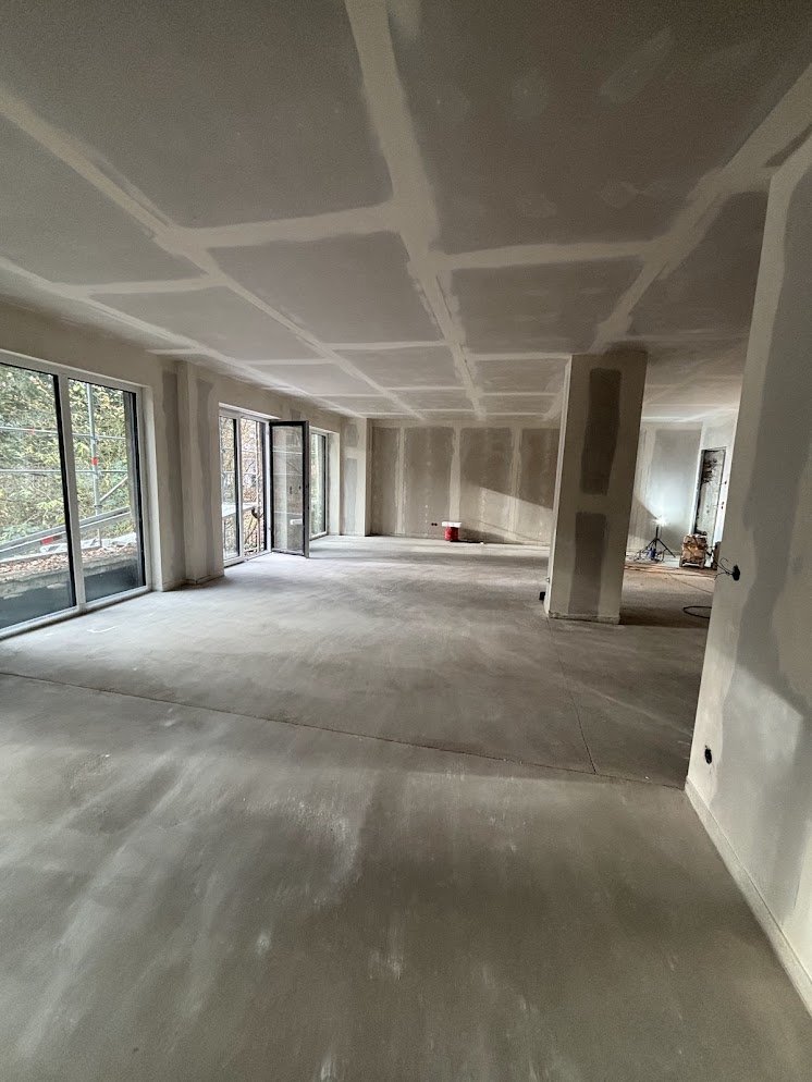 Un finished, unfurnished room in a building under construction, with large windows and visible construction materials.