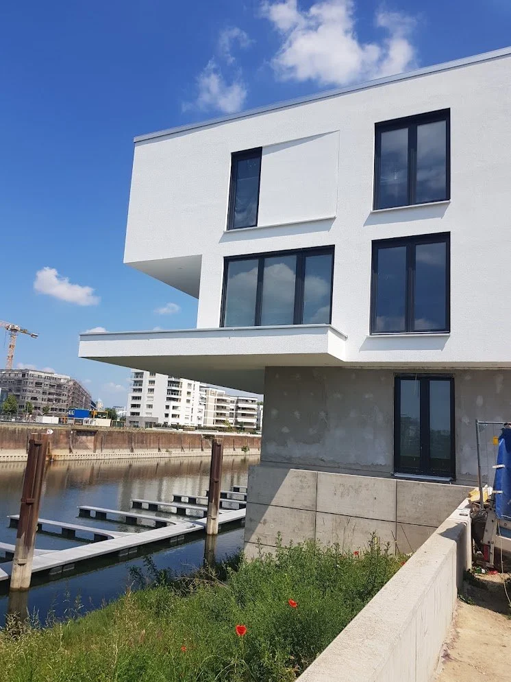 Moderner weißer Wohnblock am Wasser mit mehreren Fenstern und Balkon, Bauarbeiten im Vordergrund, blauer Himmel mit wenigen Wolken.