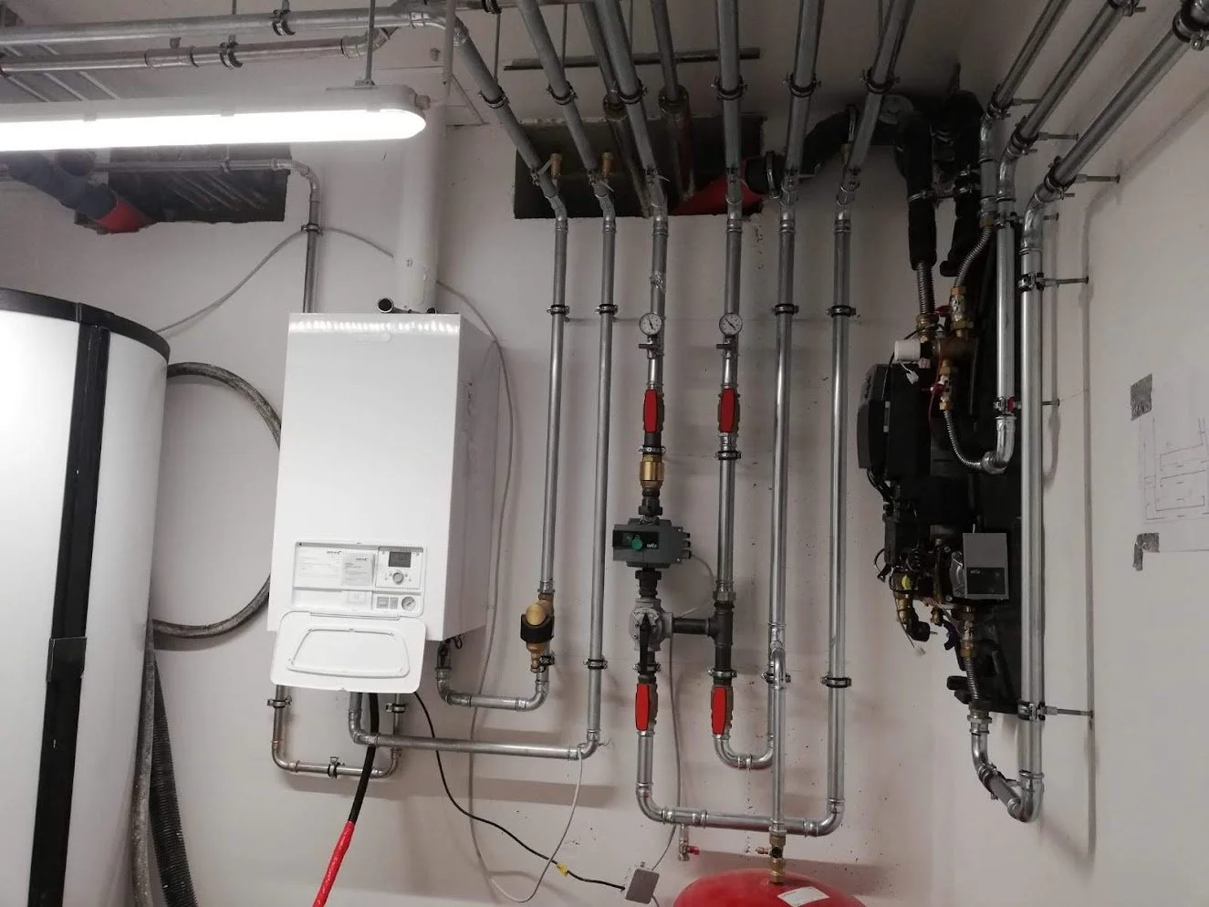 Innenaufnahme einer technischen Heizungsinstallationsanlage mit Rohren, Druckanzeigen und Steuerelementen an der Wand.