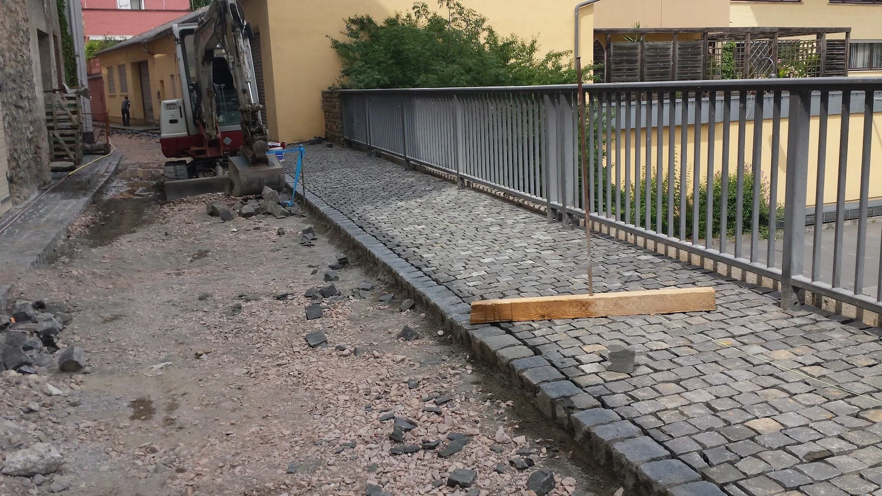 Bauarbeiten auf Gehweg, mit Baugerät, Steinen und Holzlatte, die als Wasserwaage dient, während an der Pflasterung gearbeitet wird.