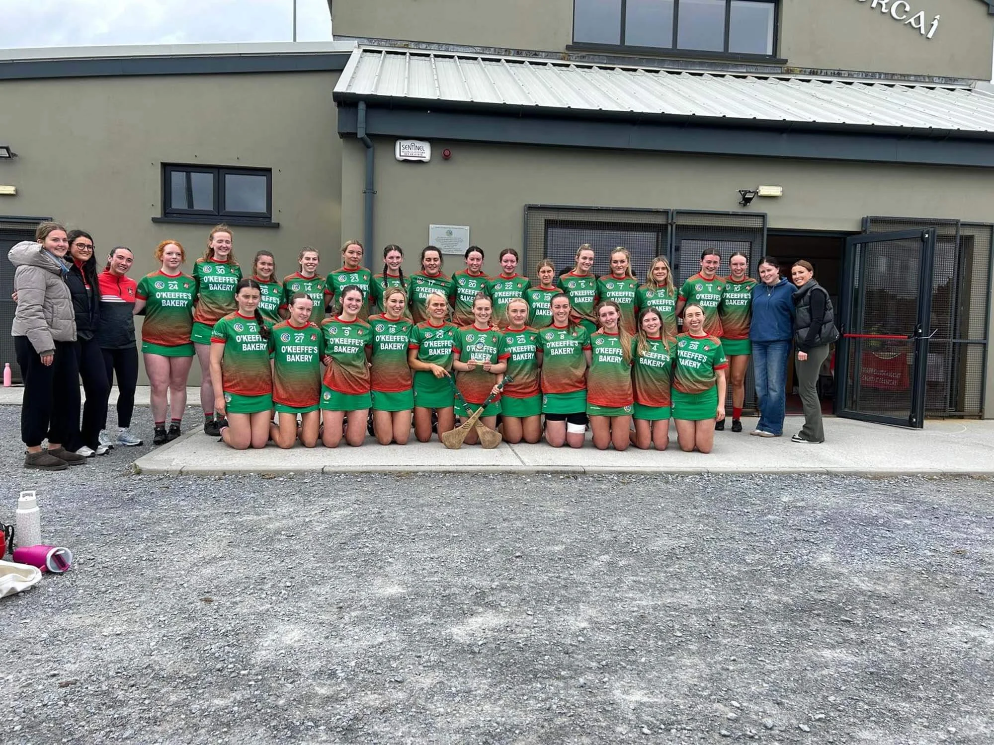 A group of women in green sports uniforms with red and brown accents, posing for a photo outside a building. Some women are standing, others kneeling, with a few people standing to the left and right. The building behind them has metal fencing and a sign that reads 'Sentinel'.
