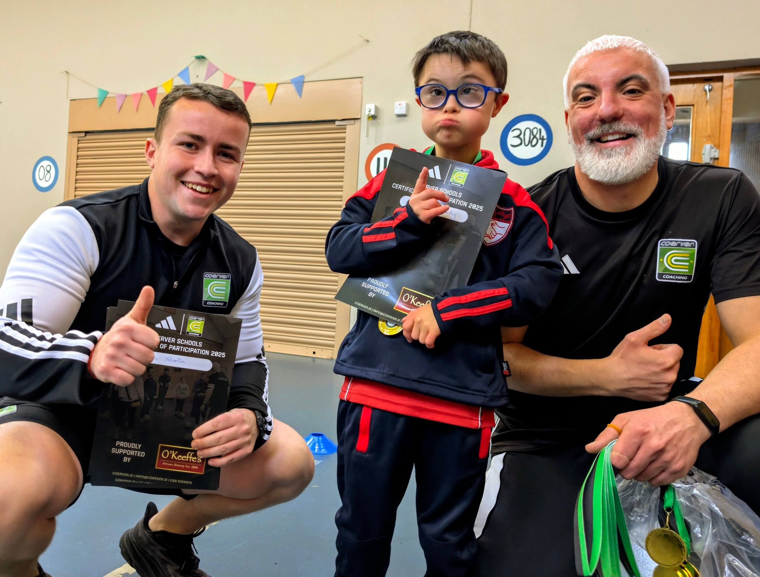 Young participant holding an O’Keeffe’s Bakery-sponsored certificate alongside two coaches at the OLGC Schools programme.