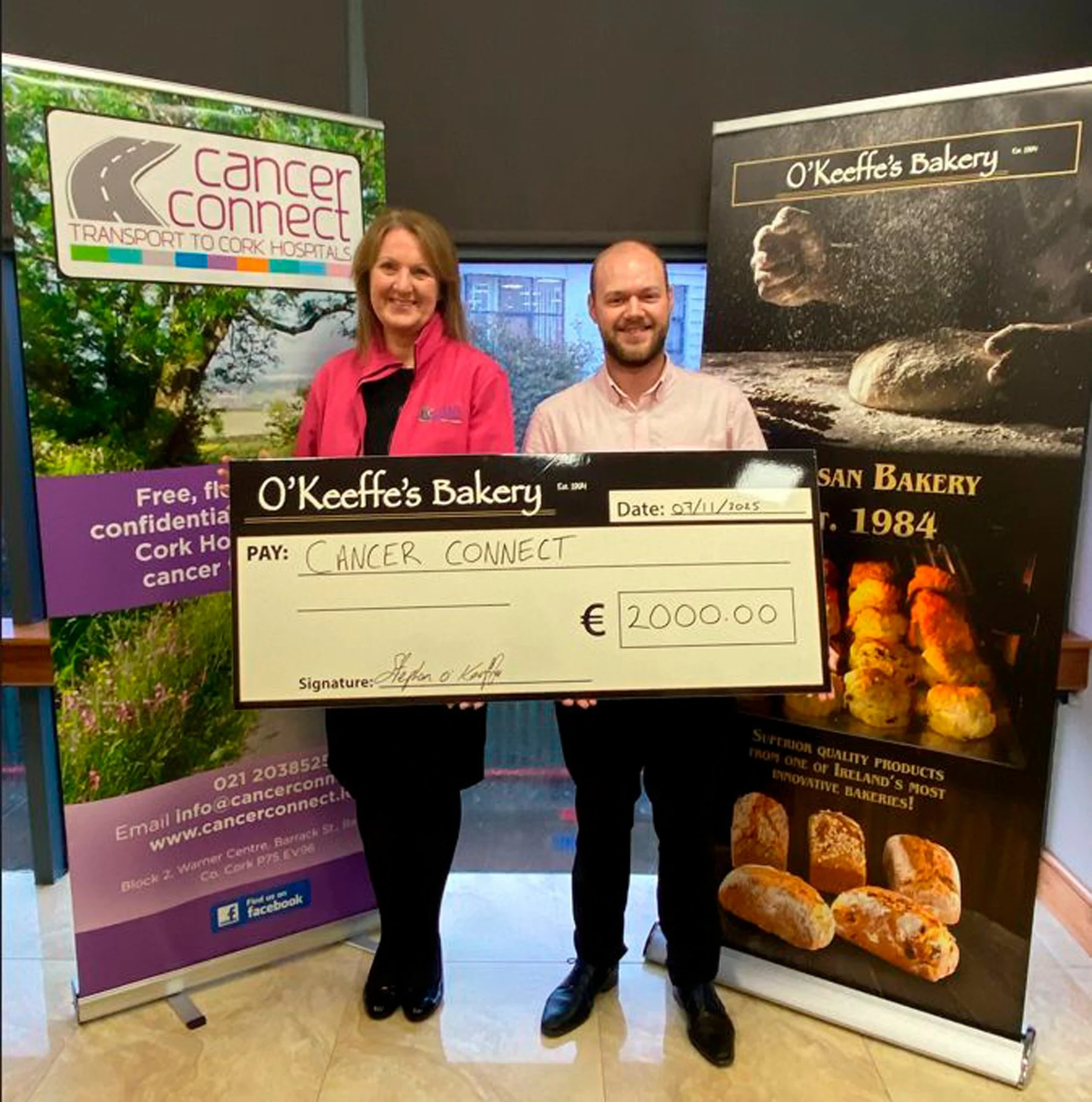 Two people holding a large cheque for €2,000 made out to Cancer Connect, standing between Cancer Connect and O'Keeffe's Bakery banner stands