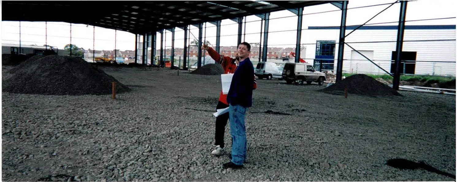 Two men standing inside a large, partially constructed building with a dirt floor, metal framing, and open sides, with parked vehicles and buildings visible outside.