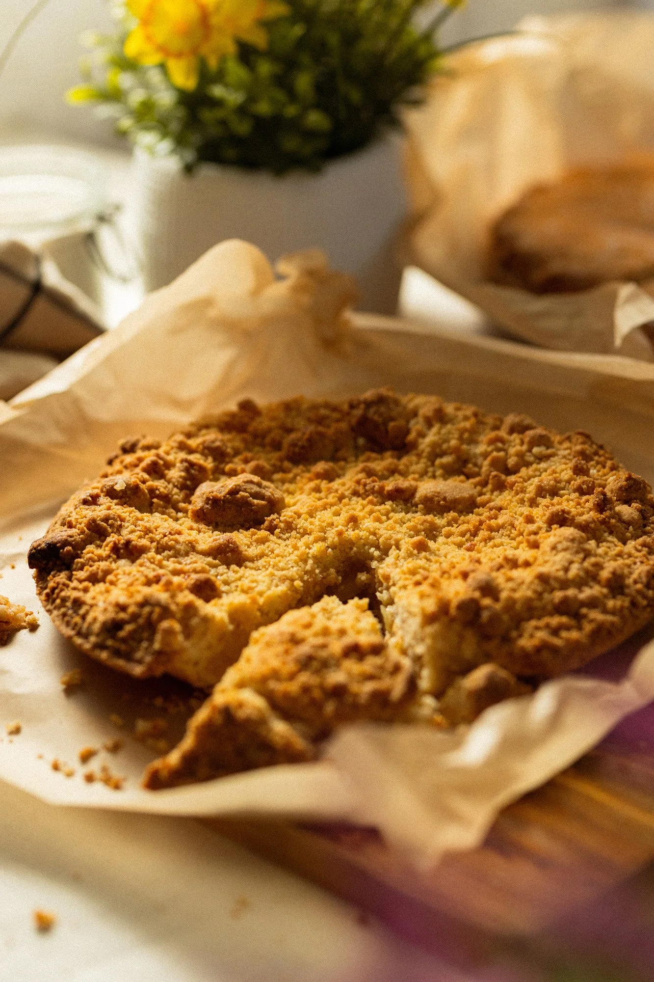 Freshly baked crumb-topped tart on baking paper with a slice removed, displayed on a wooden serving board.
