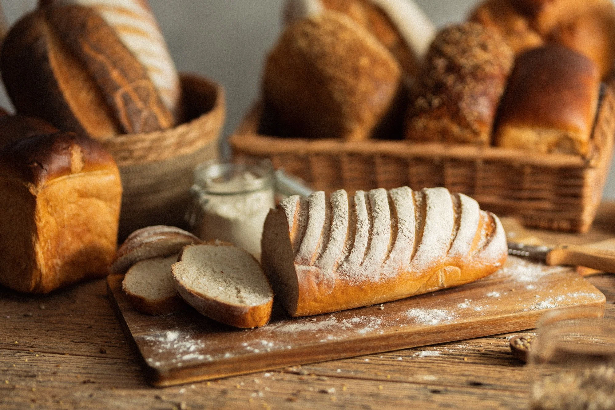 Sliced white bloomer loaf on a floured board, with a jar of milk and wheat rolls in baskets behind