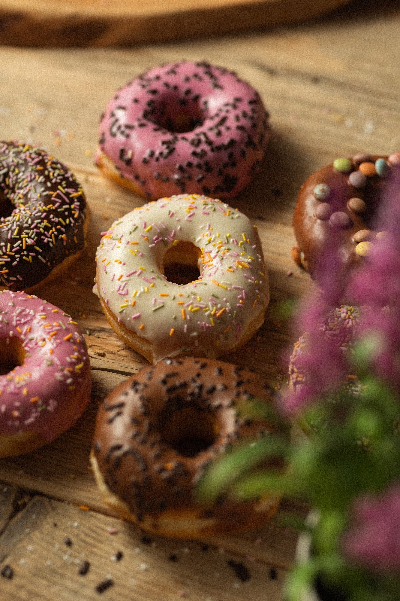 Assortment of freshly iced doughnuts with chocolate, vanilla and strawberry glazes displayed on a rustic wooden table.