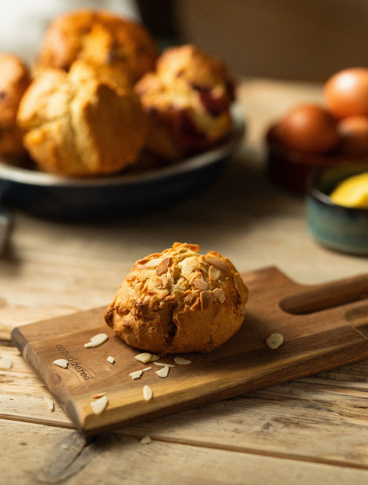 Flaked almond scone on a wooden board, shot at an angle with a bowl of scones and fresh eggs blurred behind