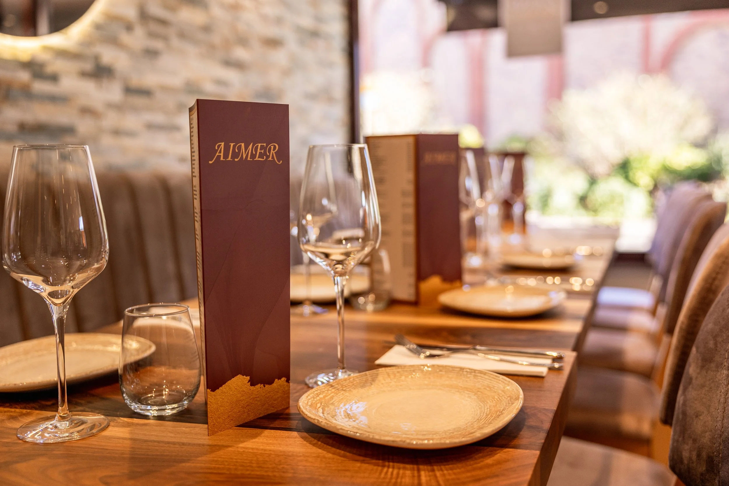 A dining table set with wine glasses, a menu, plates, and silverware in a restaurant with a brick wall and large windows.