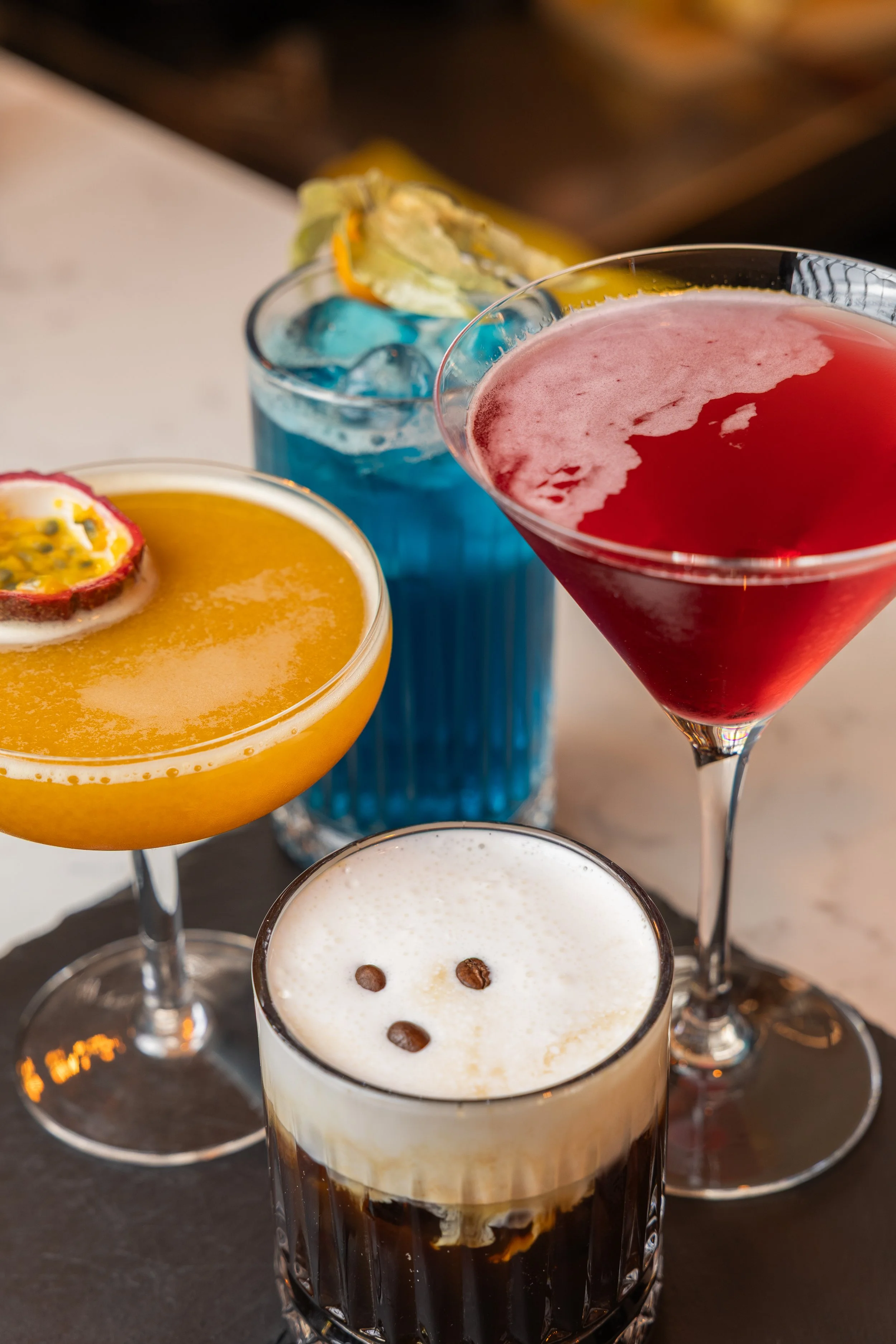 Various colorful cocktails and drinks on a table, including a dark stout with foam, a cappuccino, a passion fruit cocktail with passion fruit garnish, a blue cocktail with ice, and a pink martini. The drinks are in different types of glassware.
