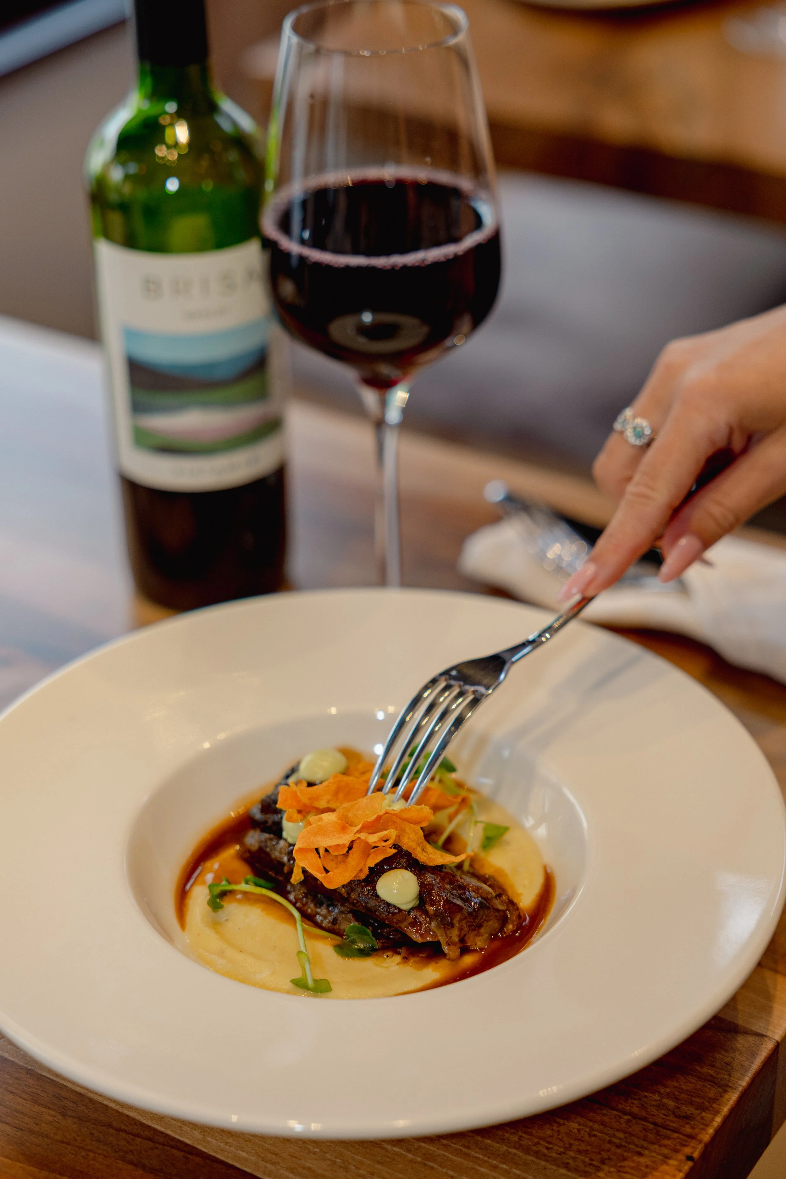 A plate of gourmet food with a piece of meat, garnished with crispy vegetables and microgreens, served in a white bowl. In the background, there is a glass of red wine and a bottle of wine.