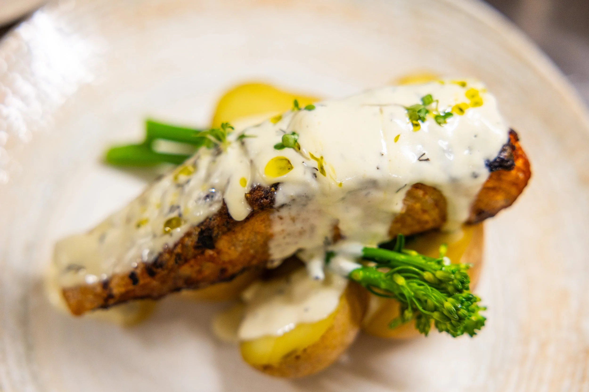 A close-up of a gourmet dish featuring a piece of cooked fish topped with a creamy white sauce, garnished with green herbs and accompanied by small vegetables and greens.
