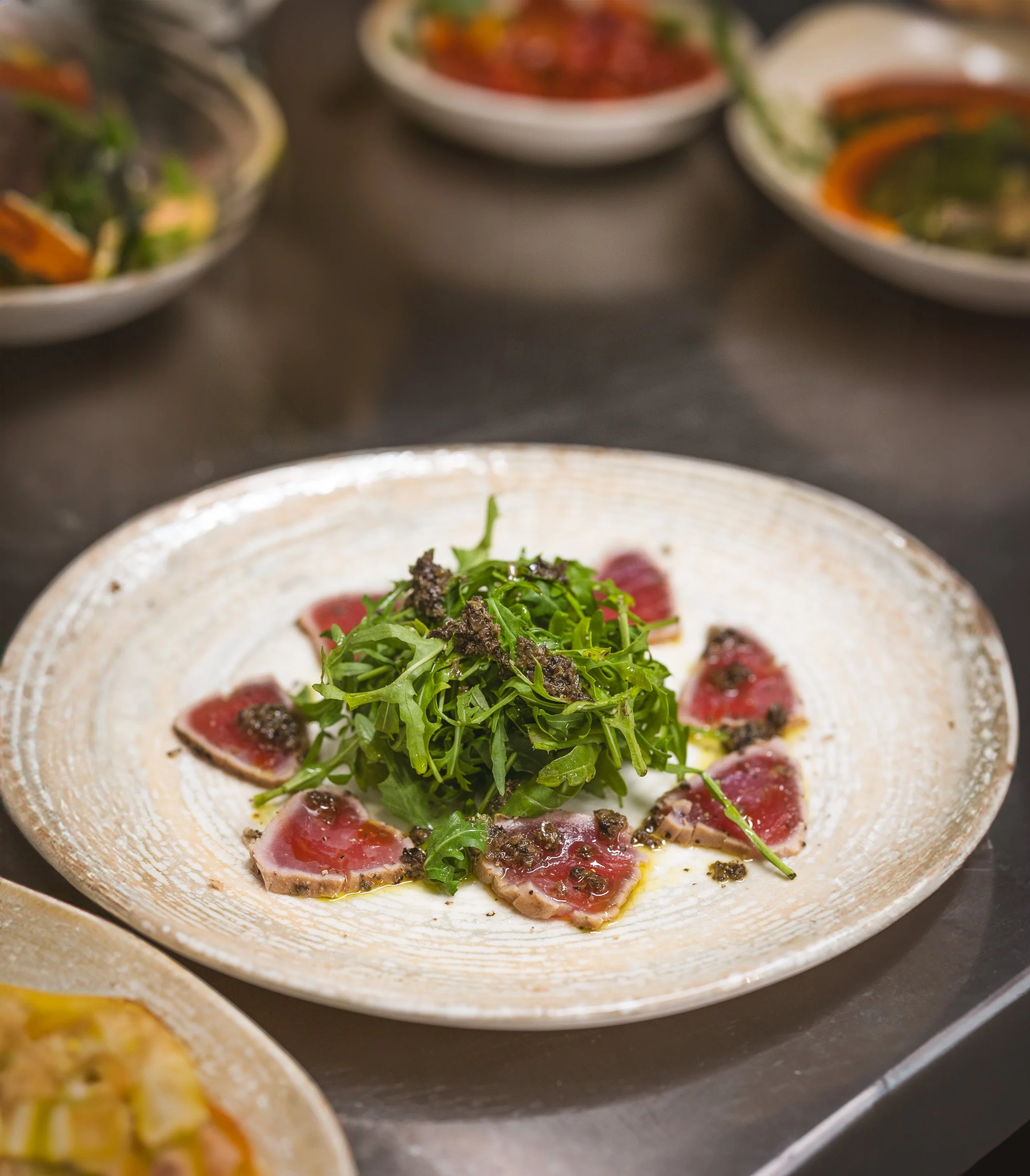 A white ceramic plate with slices of raw tuna sashimi, topped with green herbs and black sesame seeds, garnished with a small amount of oil.