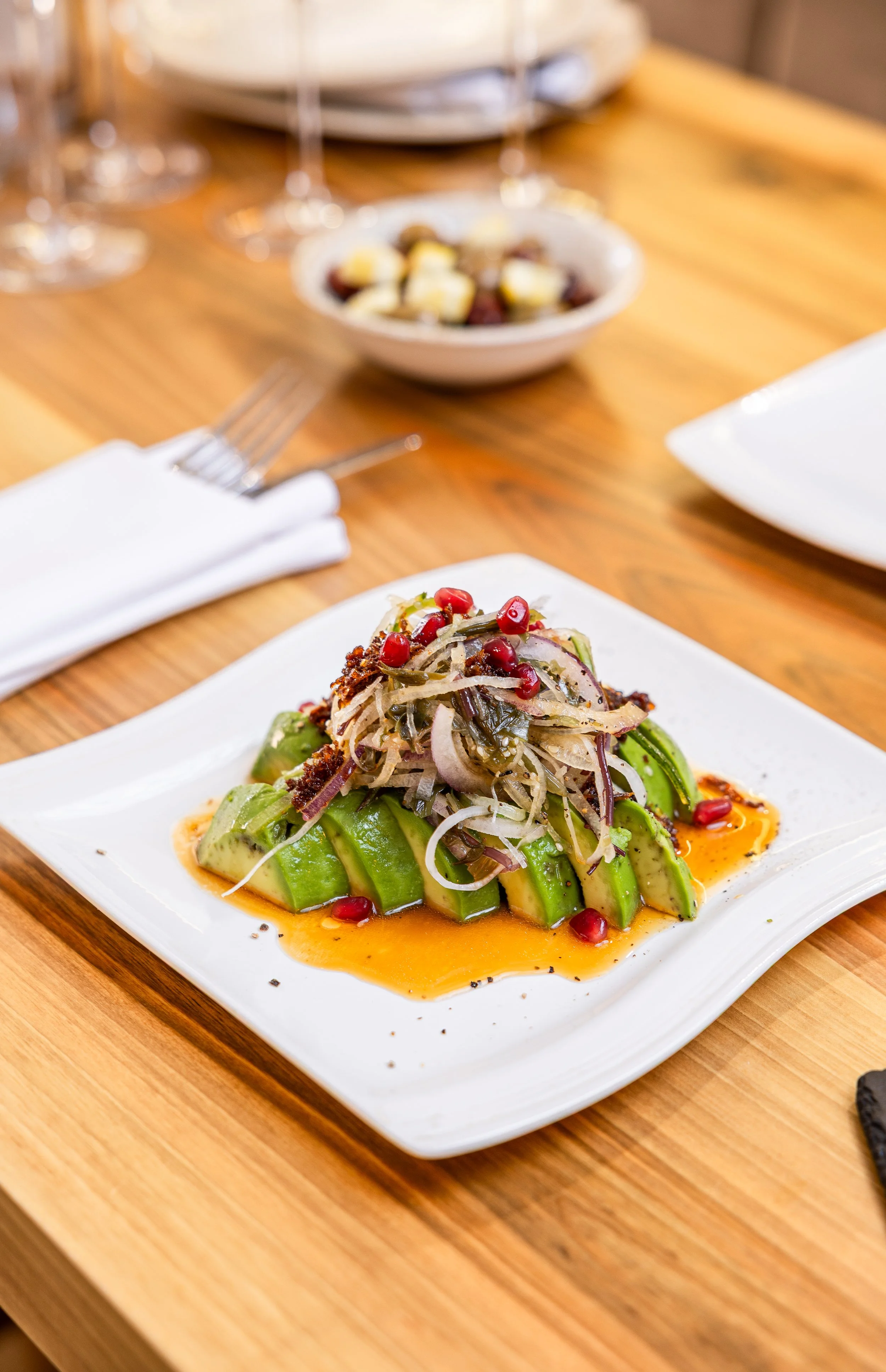 A plated dish with sliced avocado topped with a mix of vegetables and pomegranate seeds, served with a sauce on a white square plate on a wooden table.