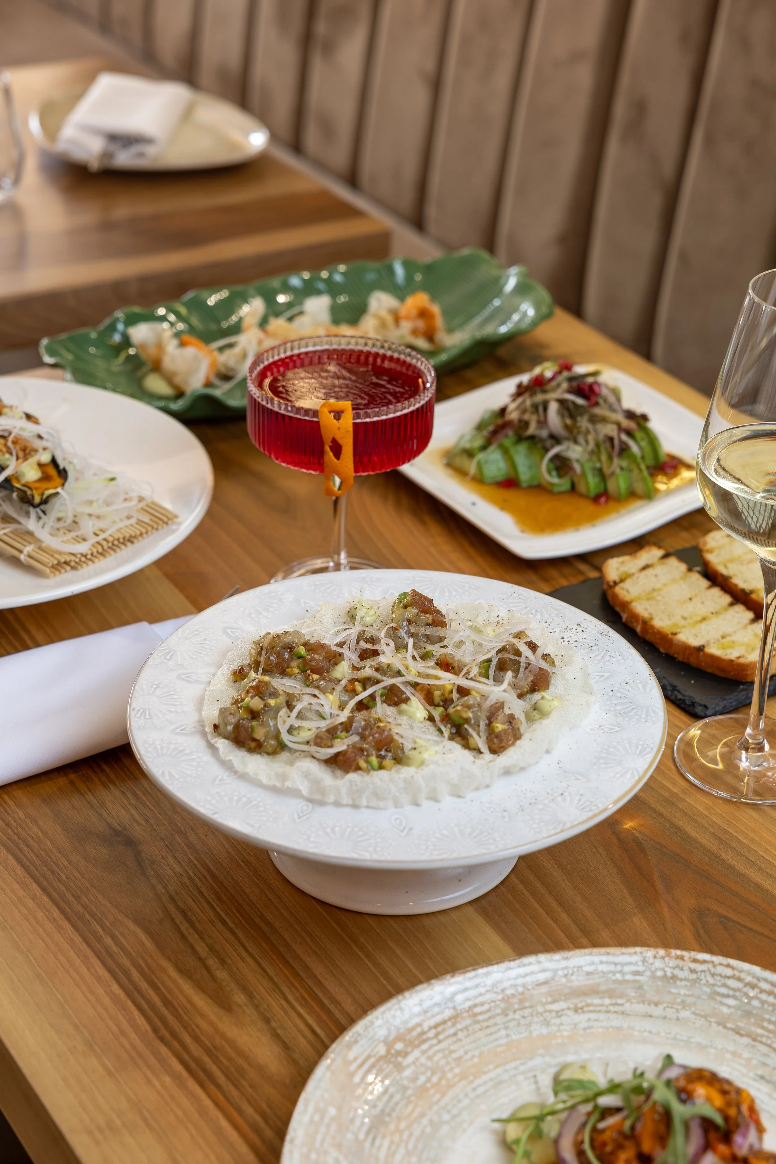 Variety of Asian dishes on white, green, and black plates on a wooden table, including a taco with meat, an arranged salad, dumplings, and a glass of red beverage.
