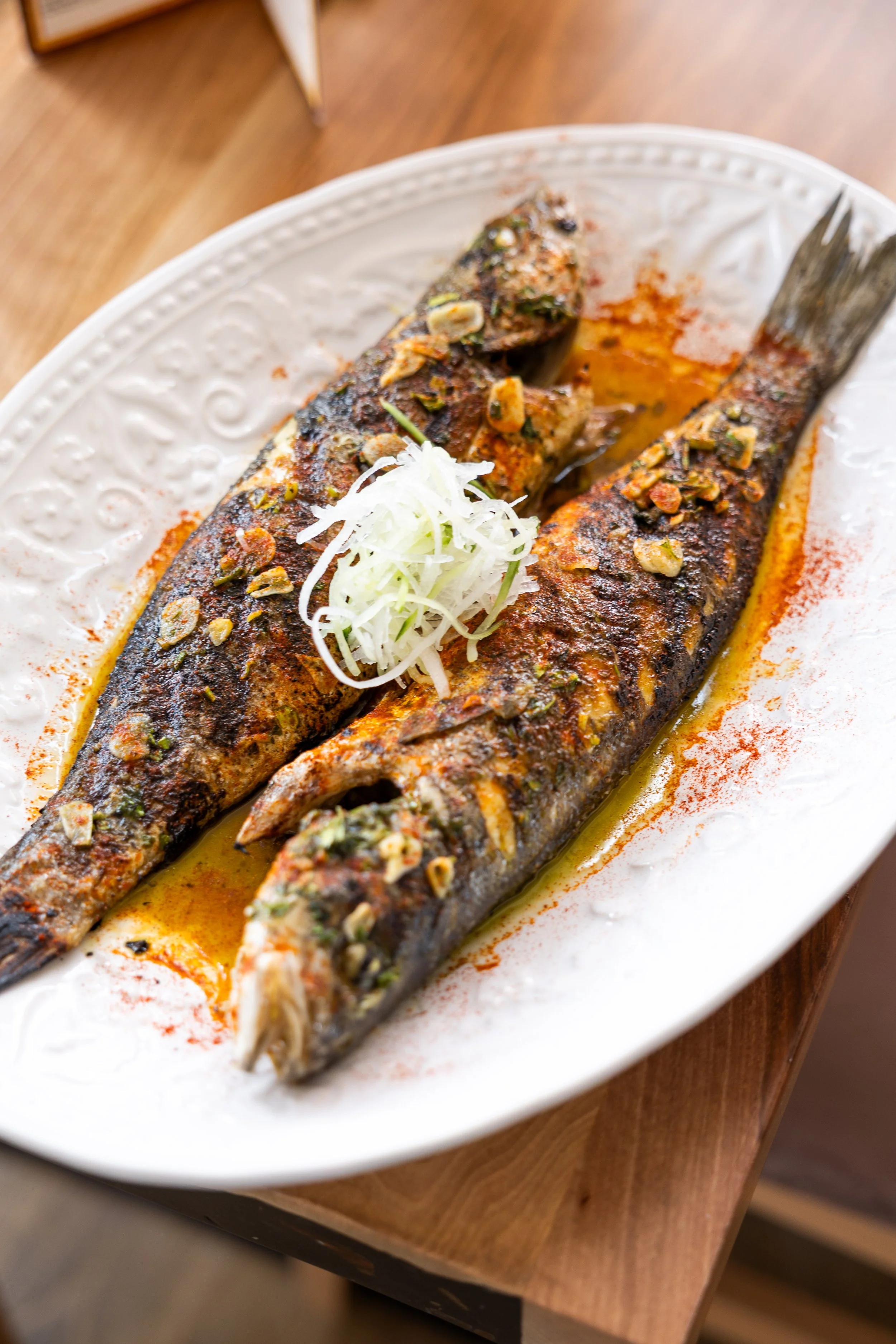 Grilled whole fish topped with chopped herbs and onions on a white decorative plate with a yellow sauce and red paprika powder, placed on a wooden table.