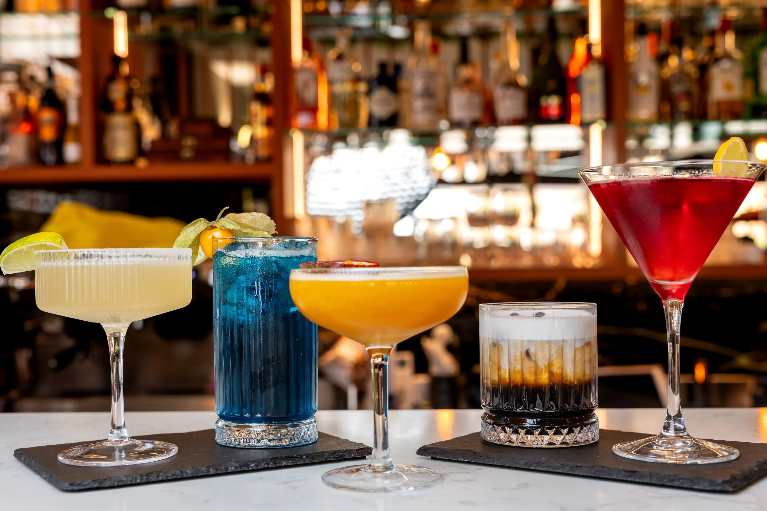 An assortment of colorful cocktails on a bar counter in front of a blurred bar background.
