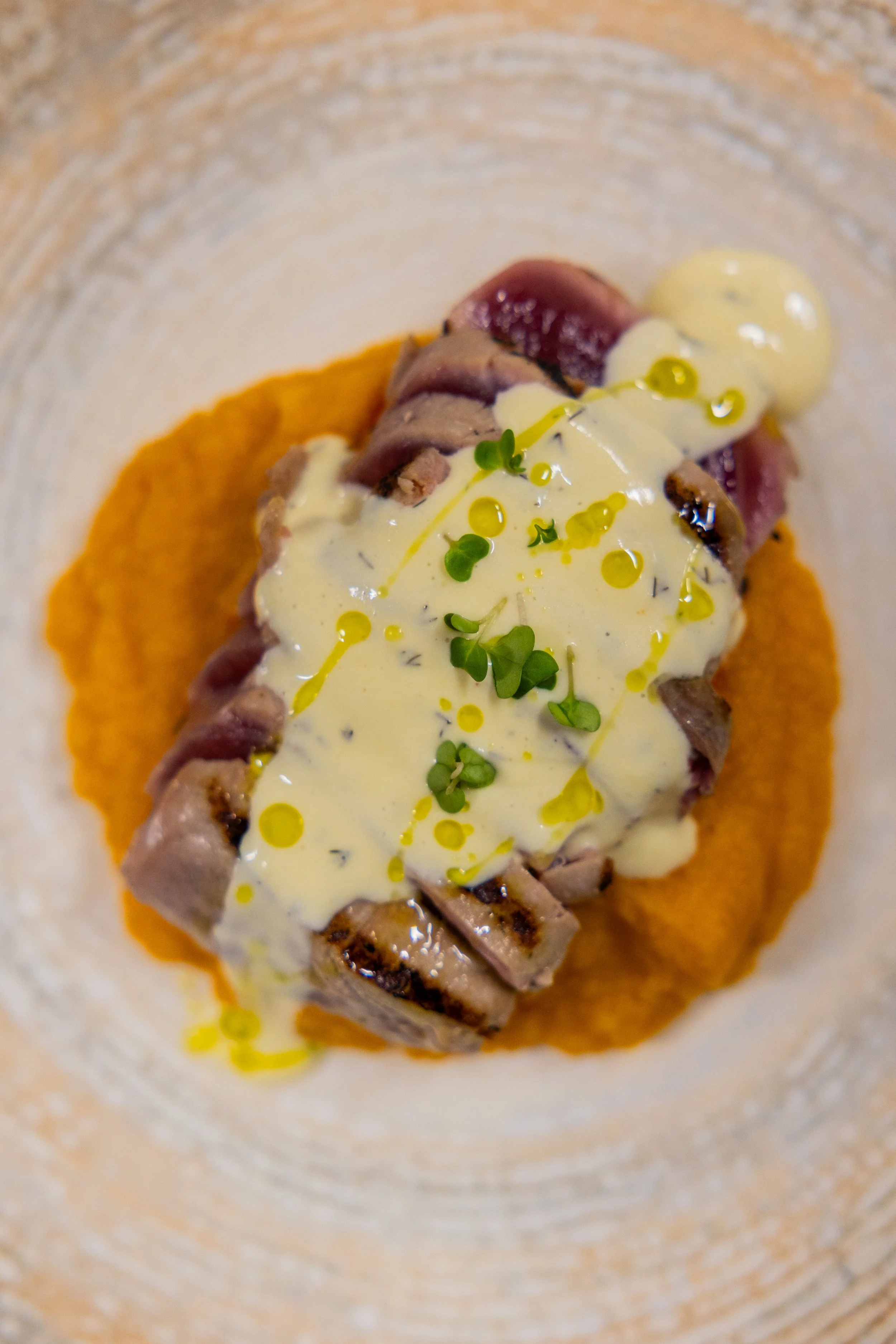 Close-up of sliced roasted meat topped with creamy sauce, microgreens, and olive oil, plated on an orange puree.