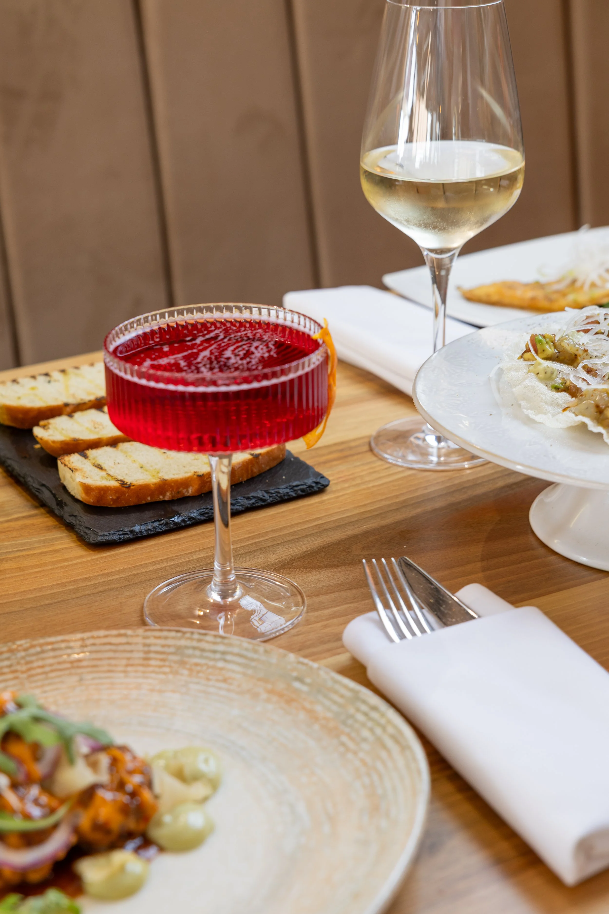 A table setting with a glass of white wine, a glass of red wine, a plate of food, and bread with a butter knife on a white napkin.