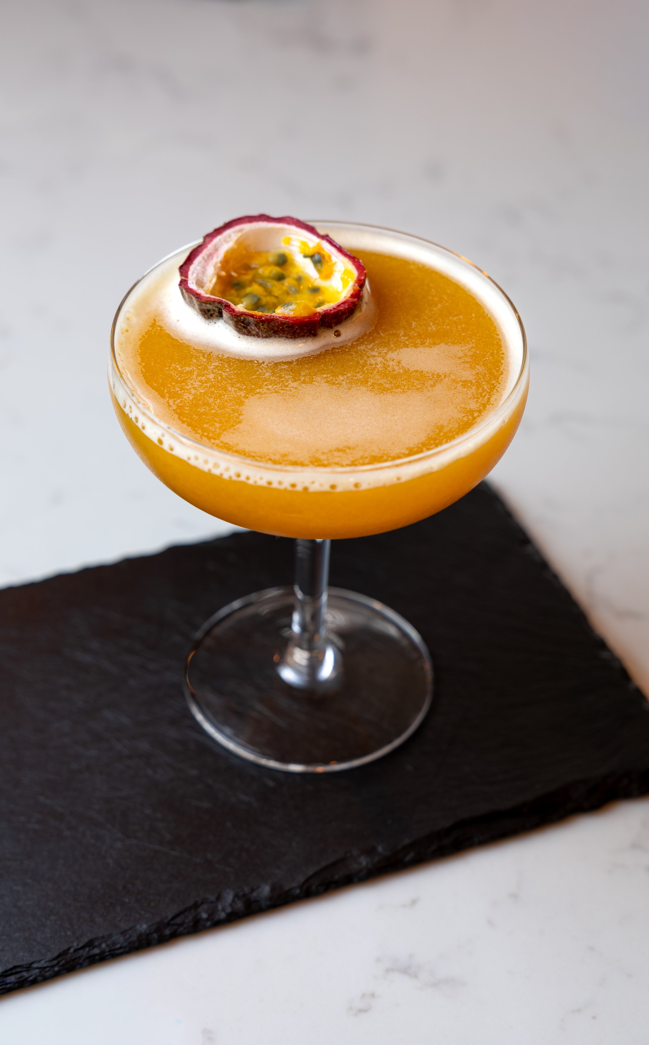 A cocktail in a coupe glass garnished with a passion fruit half.