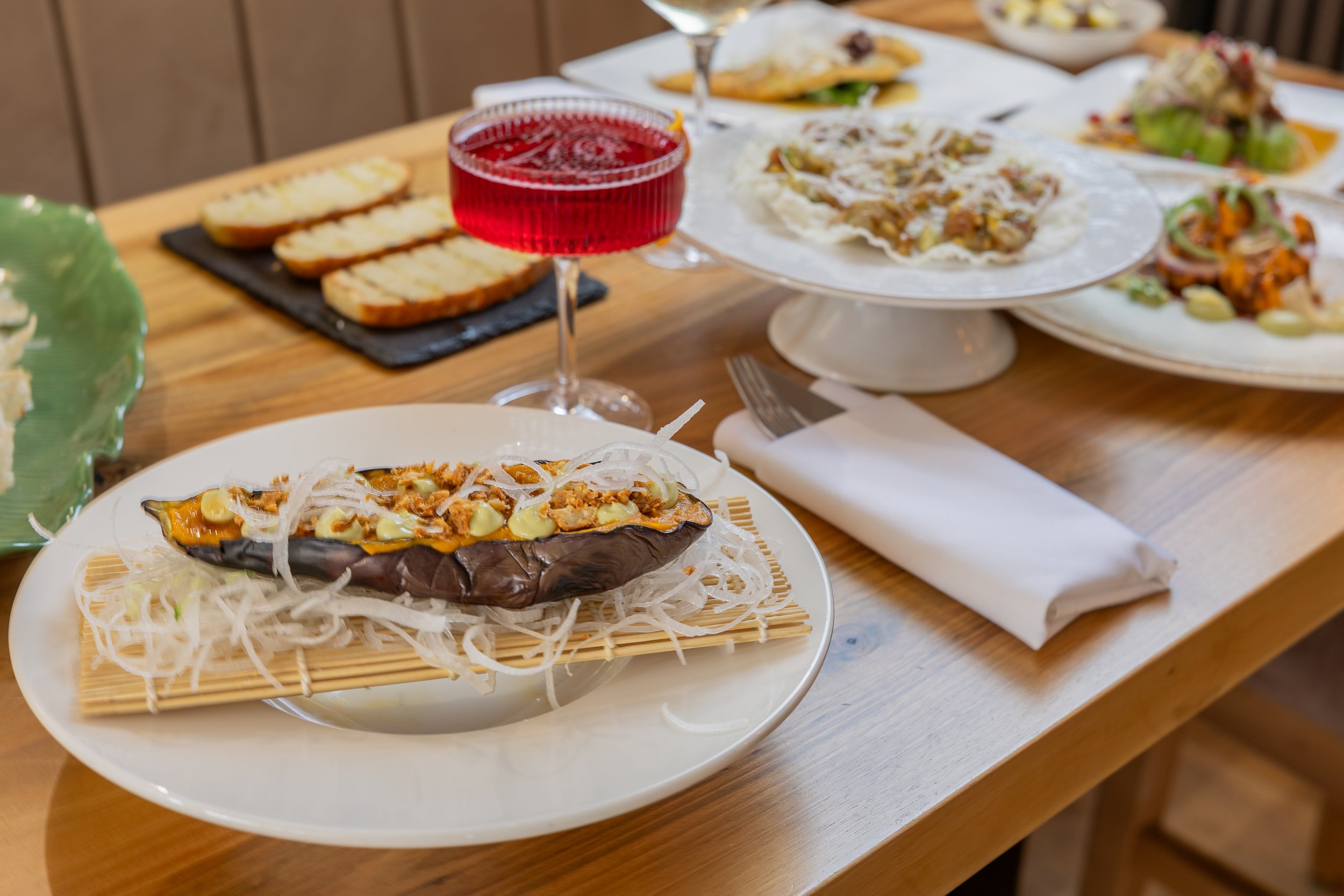 A wooden table with various dishes including stuffed eggplant on a bed of shredded white vegetables on a white plate, slices of bread on a black slate, and several plates of tacos with various toppings. A glass of red beverage is in focus in the cent